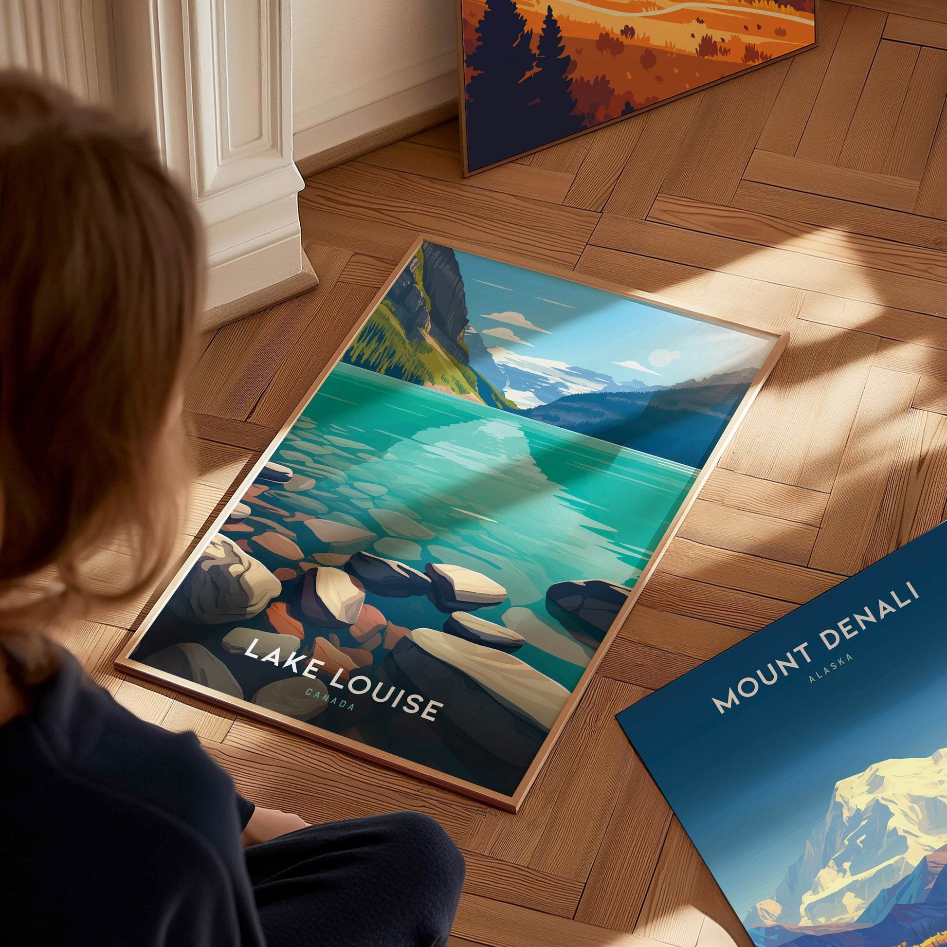 A person observes a Lake Louise poster featuring turquoise waters and rocky shores, placed on a wooden floor near a Mount Denali poster. Sunlight casts shadows across the scene.