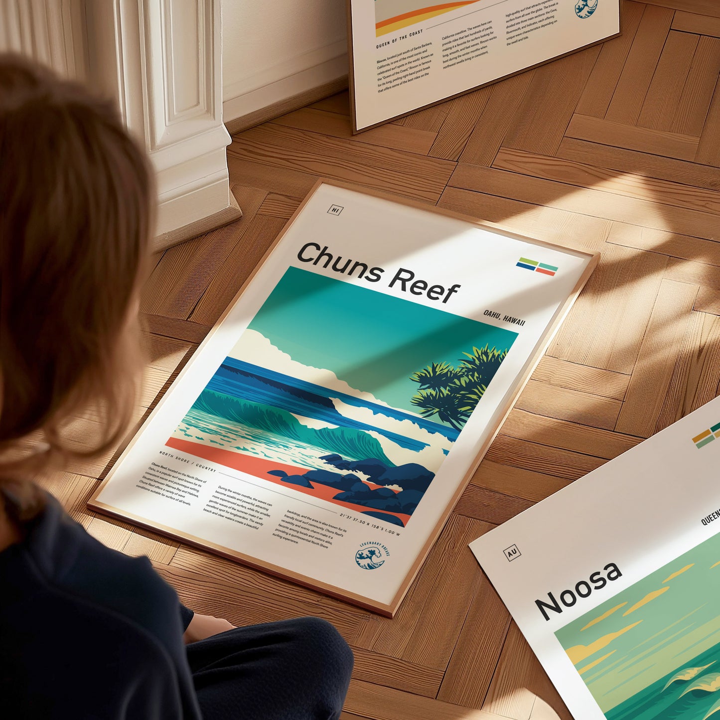 A child sits on a wooden floor, looking at a framed poster titled Chuns Reef. The vibrant illustration features rolling waves and lush greenery, capturing the essence of Oahus North Shore surf culture.