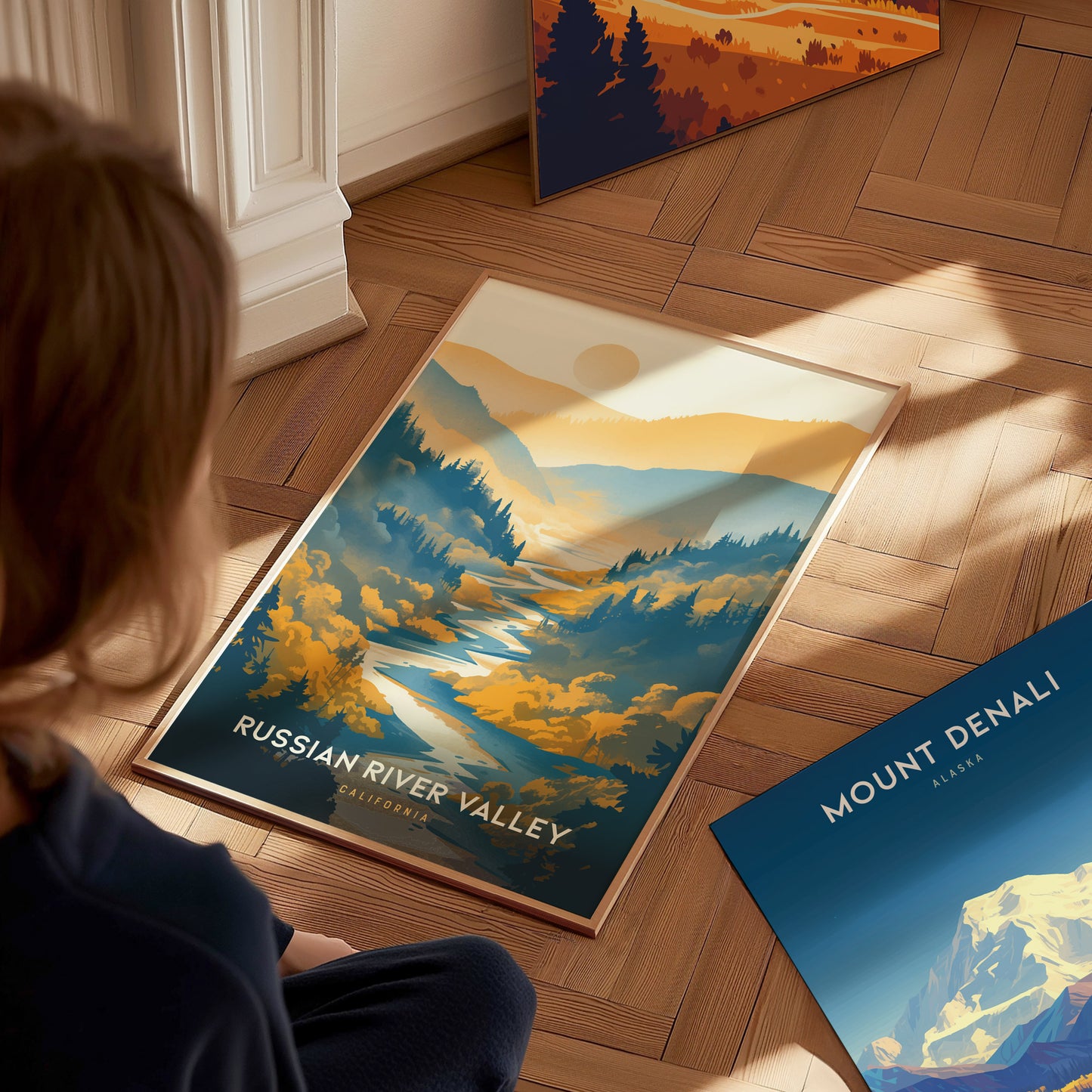 A person looks at a vibrant poster of the Russian River Valley on a wooden floor, featuring colorful landscapes and a sunlit sky.