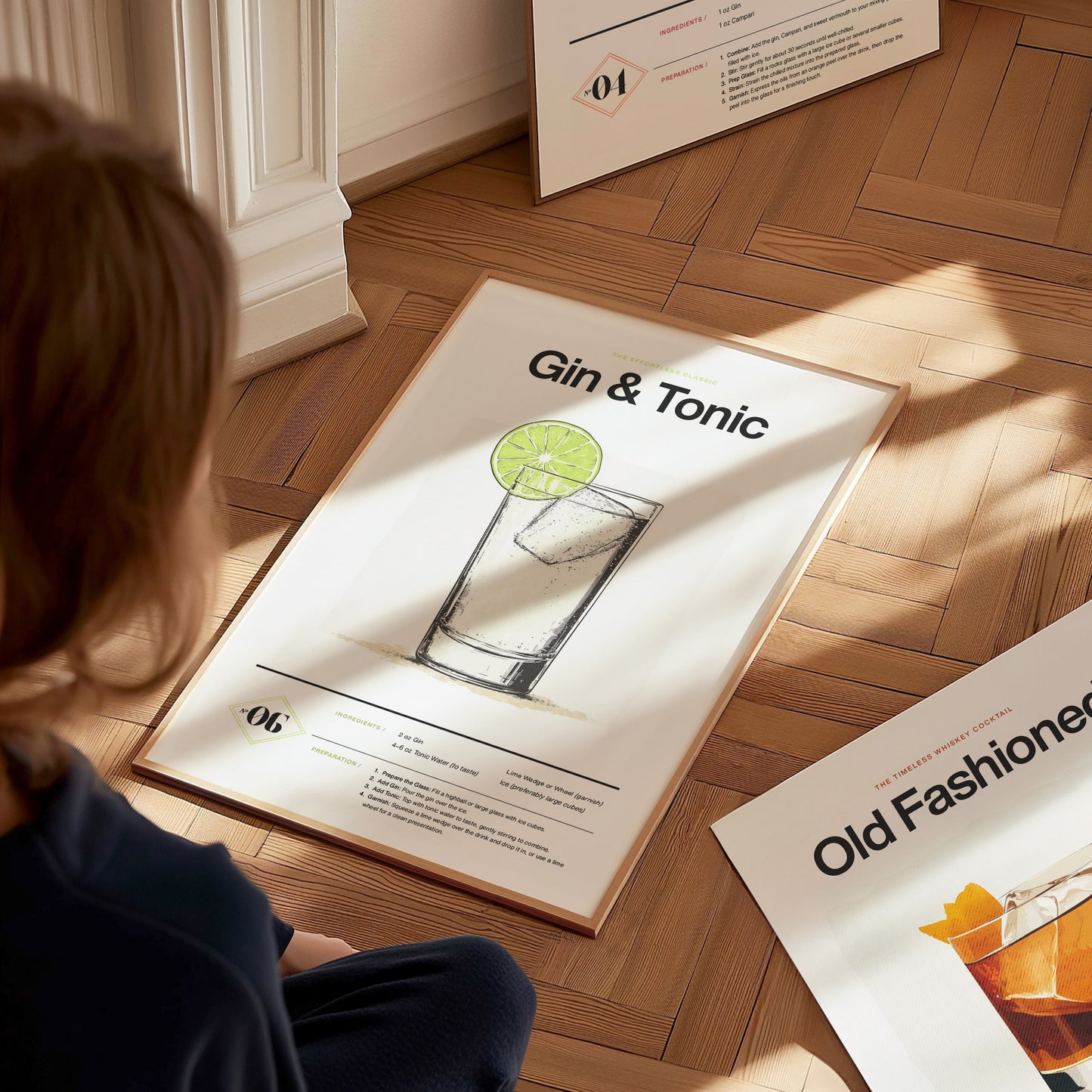 A person looks at a framed Gin and Tonic cocktail poster on a wooden floor, featuring a minimalist design with a lime garnish illustration. Additional posters are partially visible nearby.