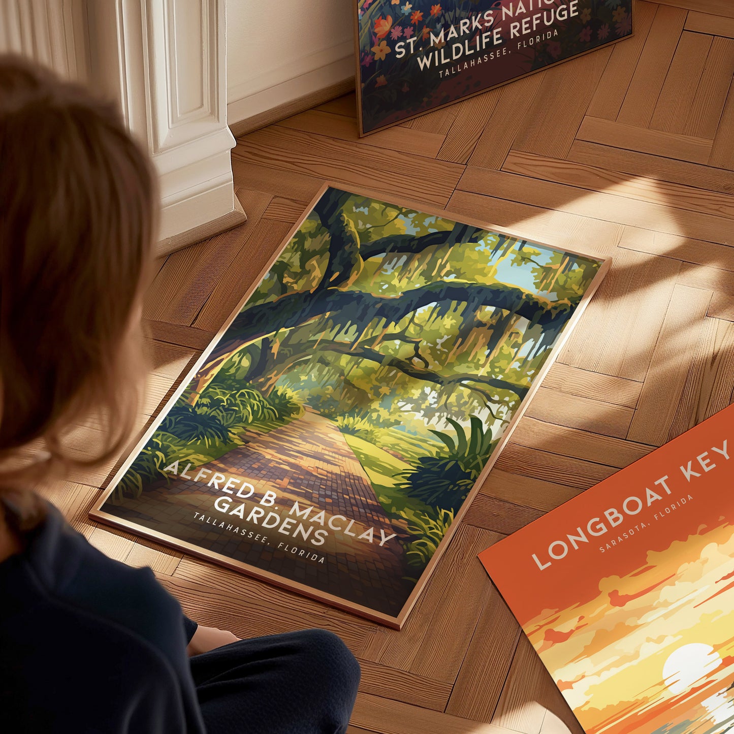 A person sits on a wooden floor gazing at a vibrant poster of Alfred B. Maclay Gardens, featuring a scenic path under moss-draped trees. Other posters are partially visible nearby.