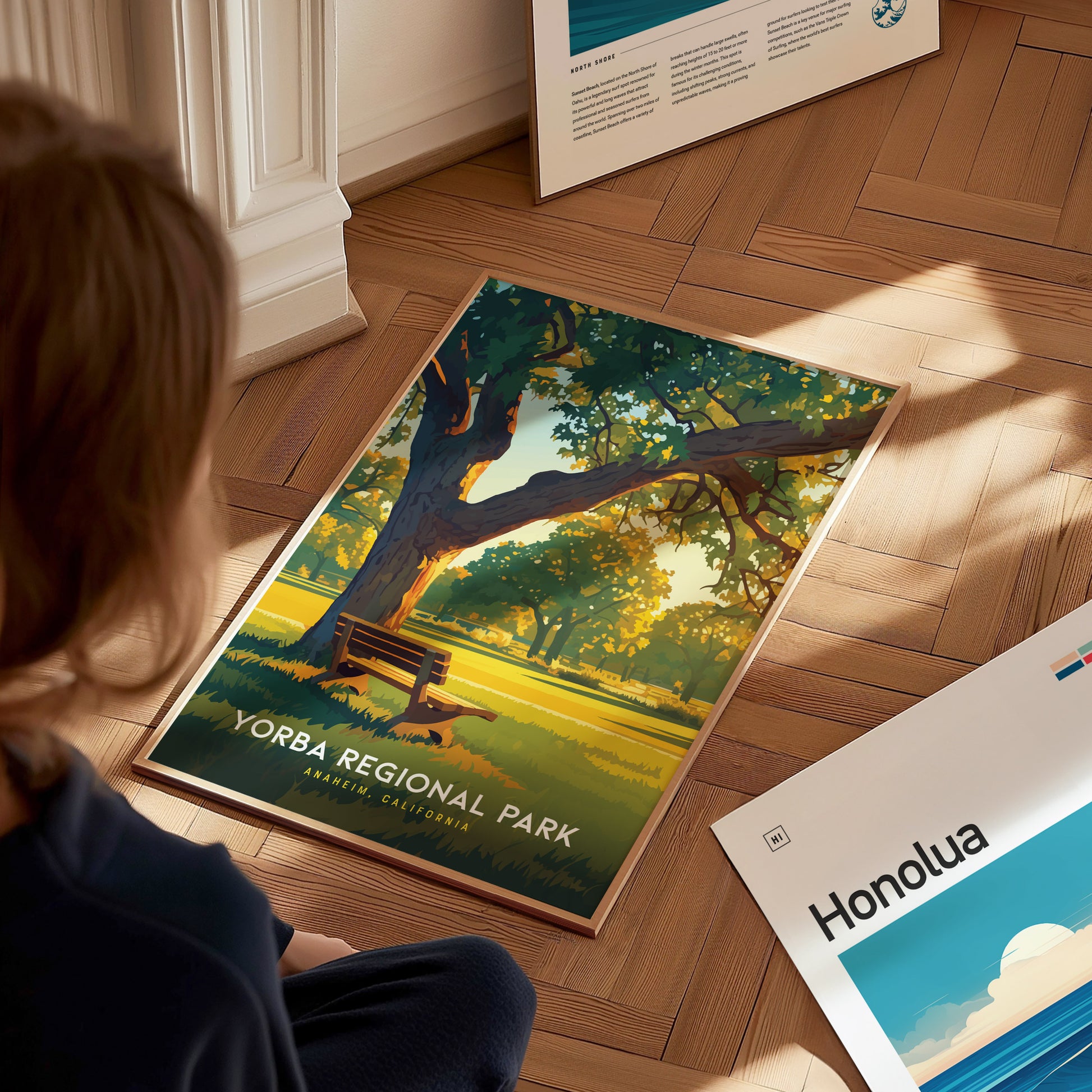 A person observes a Yorba Regional Park poster on a wooden floor, depicting a sunlit park scene with a bench under a tree. Other posters are partially visible nearby.