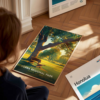 A person observes a Yorba Regional Park poster on a wooden floor, depicting a sunlit park scene with a bench under a tree. Other posters are partially visible nearby.