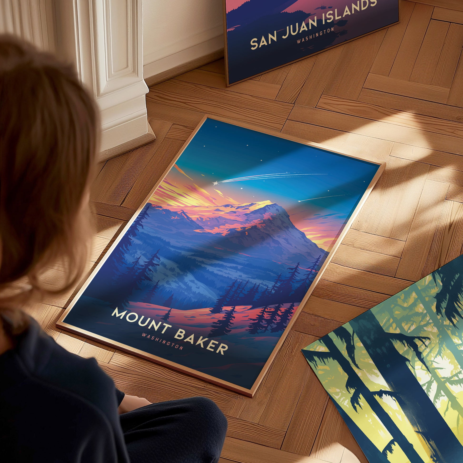 A person gazes at a vibrant Mount Baker poster on a wooden floor, depicting a colorful sunset over a mountainous landscape. Nearby, another poster titled San Juan Islands is partially.
