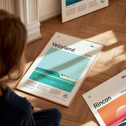 A person is sitting on the floor looking at a framed Velzyland surf poster, showcasing turquoise waves and minimalist design. Nearby, another poster titled Rincon is visible.