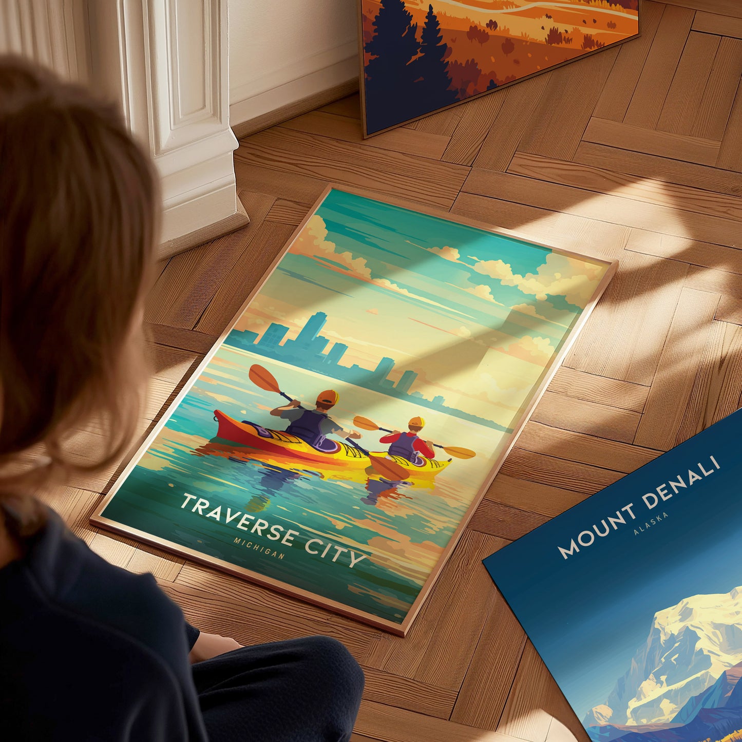 A person looks at a Traverse City poster featuring two kayakers on calm waters, with a city skyline in the background, placed on a wooden floor.