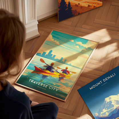 A person looks at a Traverse City poster featuring two kayakers on calm waters, with a city skyline in the background, placed on a wooden floor.
