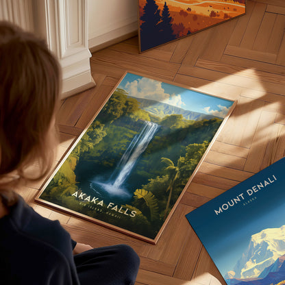 A person observes a vibrant Akaka Falls poster on a wooden floor, showcasing a waterfall amidst lush greenery. Nearby, a Mount Denali poster rests against the wall.