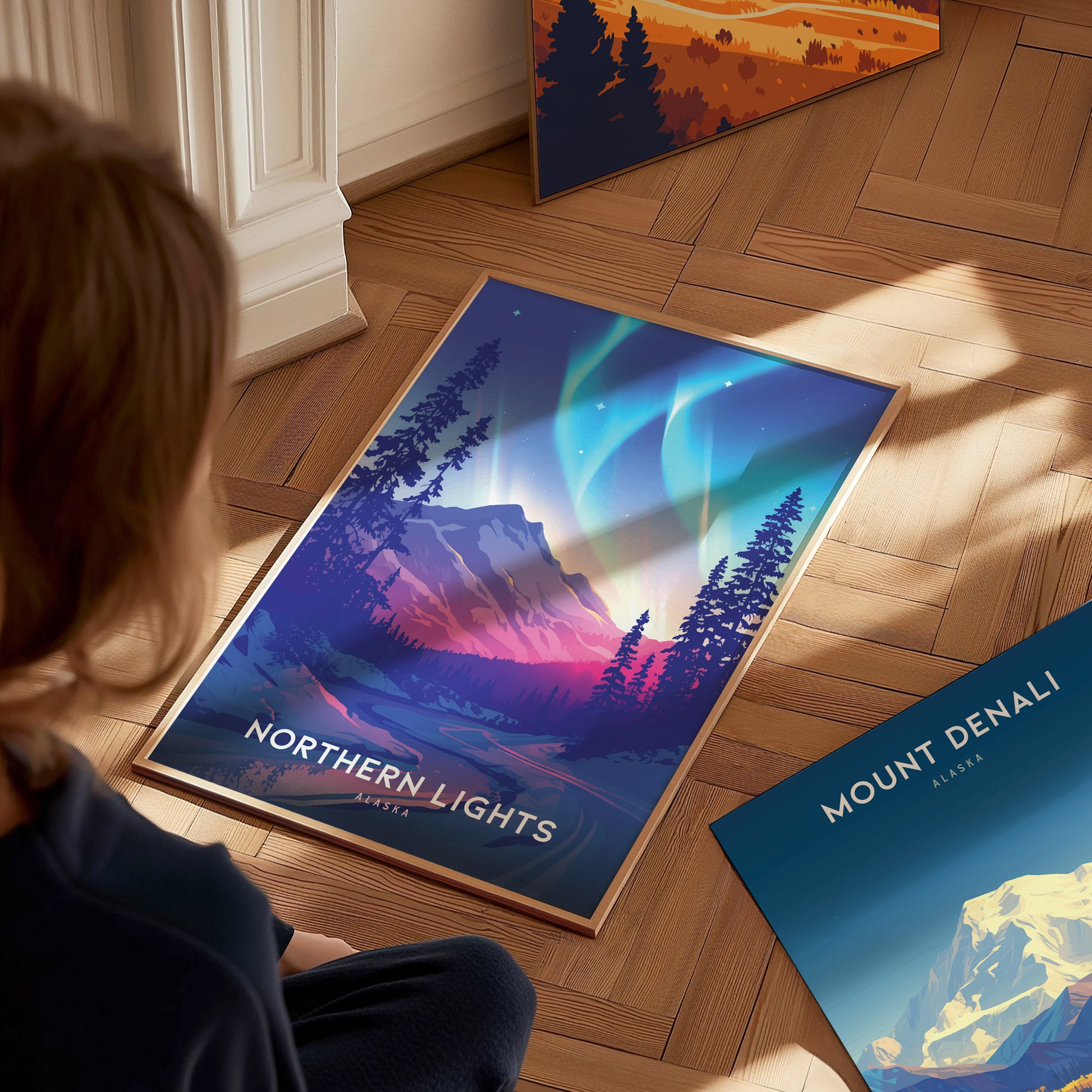 A person is looking at a vibrant poster of the Northern Lights over a mountain landscape, with another poster of Mount Denali nearby on a wooden floor.