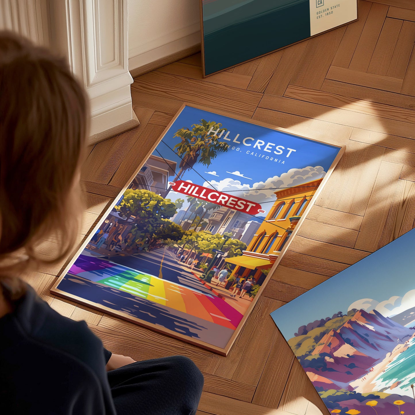 A person sits on a wooden floor, admiring a colorful poster of Hillcrest, San Diego, featuring the iconic sign and a vibrant rainbow crosswalk under a blue sky.