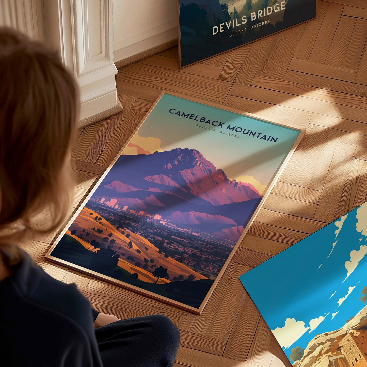 A person sits on a wooden floor, observing a vibrant Camelback Mountain poster with desert tones. Nearby, another poster depicting Devils Bridge in Sedona, Arizona is partially visible.