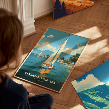 A person sits on a wooden floor, observing a framed Carmel-by-the-Sea poster featuring a sailboat on a vibrant blue sea under a bright sky. Other art pieces are nearby.
