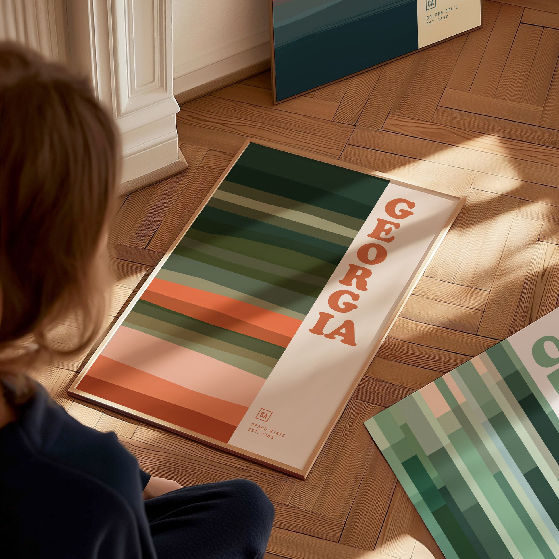 A person sits on a wooden floor, looking at a framed poster featuring bold stripes in earthy tones and the word Georgia in large letters. Sunlight casts soft shadows.