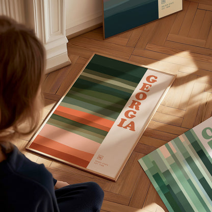A person sits on a wooden floor, looking at a framed poster featuring bold stripes in earthy tones and the word Georgia in large letters. Sunlight casts soft shadows.