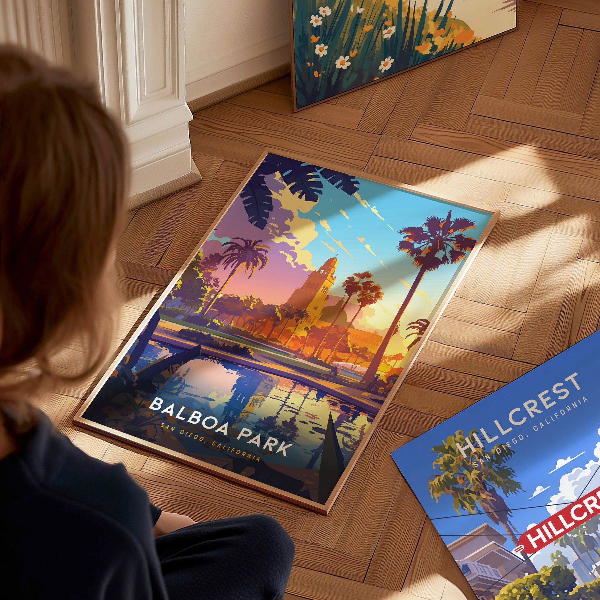 A person sits on a wooden floor gazing at a colorful Balboa Park poster depicting palm trees and vibrant skies. Nearby, another Hillcrest poster rests on the floor.