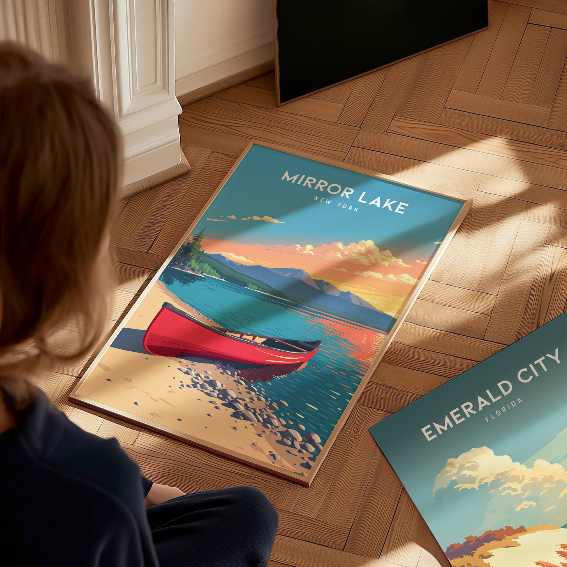 A child is sitting on a wooden floor, gazing at a colorful poster featuring Mirror Lake, New York. The artwork depicts a serene lake scene with a red canoe, vibrant mountains, and a sunset sky, capturing the tranquil beauty of.