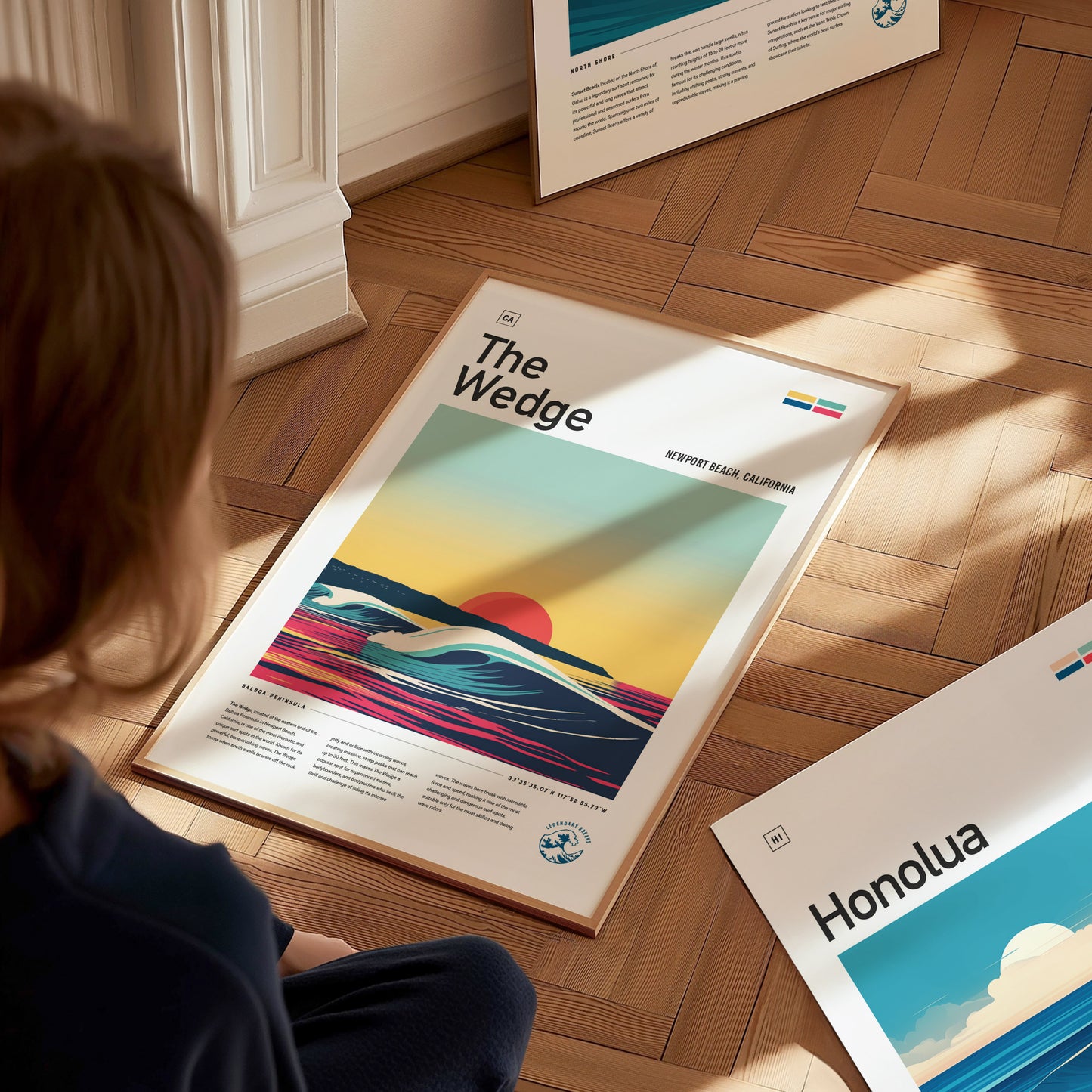 A person sits on a wooden floor admiring a framed poster titled The Wedge with a vibrant sunset and wave design. Nearby, another poster labeled Honolua is partially visible.