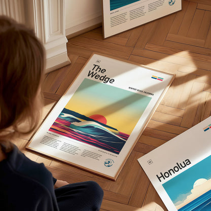 A person sits on a wooden floor admiring a framed poster titled The Wedge with a vibrant sunset and wave design. Nearby, another poster labeled Honolua is partially visible.