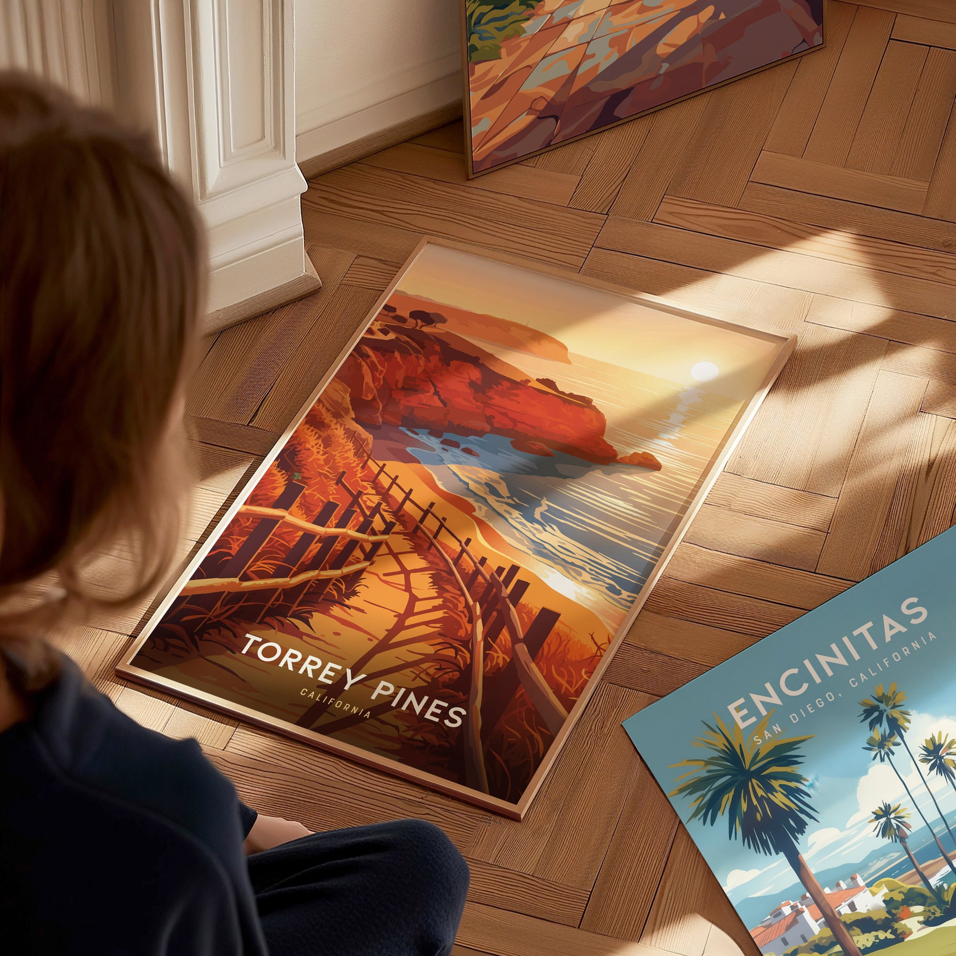 A person views a vibrant Torrey Pines poster on a wooden floor, depicting coastal cliffs and a sunset. Nearby, an Encinitas poster features palm trees and ocean scenery.