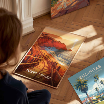 A person views a vibrant Torrey Pines poster on a wooden floor, depicting coastal cliffs and a sunset. Nearby, an Encinitas poster features palm trees and ocean scenery.