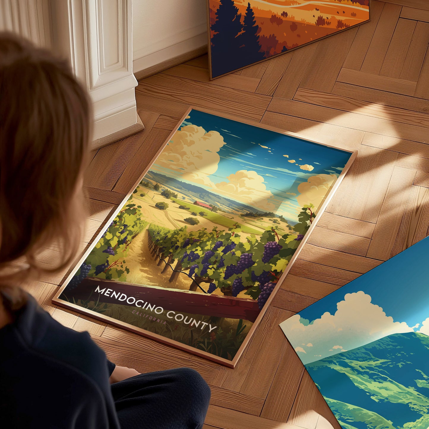 A person sits on a wooden floor, looking at a Mendocino County poster depicting vineyards and a coastal landscape. Additional posters with scenic artwork are visible nearby.