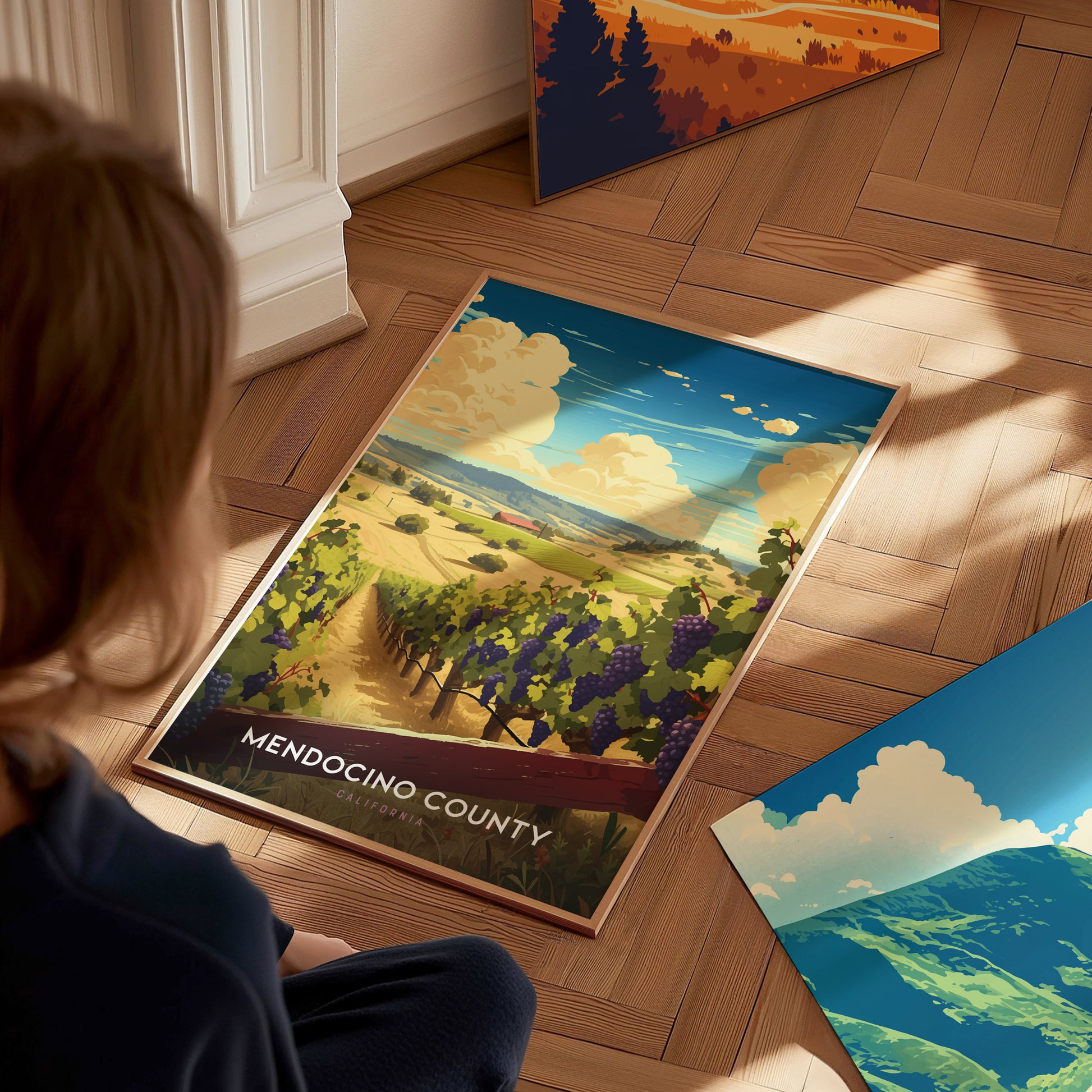 A person sits on a wooden floor, looking at a Mendocino County poster depicting vineyards and a coastal landscape. Additional posters with scenic artwork are visible nearby.
