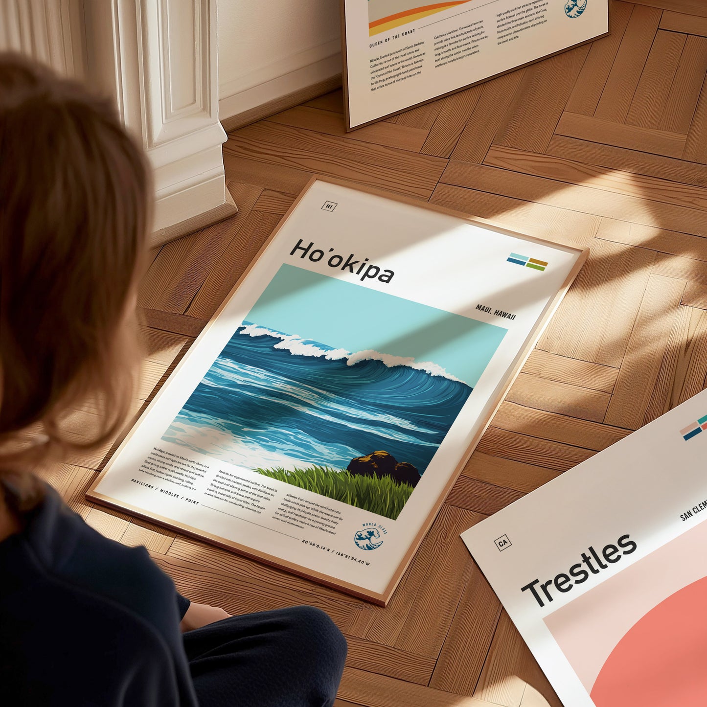 A person gazes at a framed poster of Hookipa Beach featuring a vibrant wave illustration, placed on a wooden floor alongside other framed artworks.