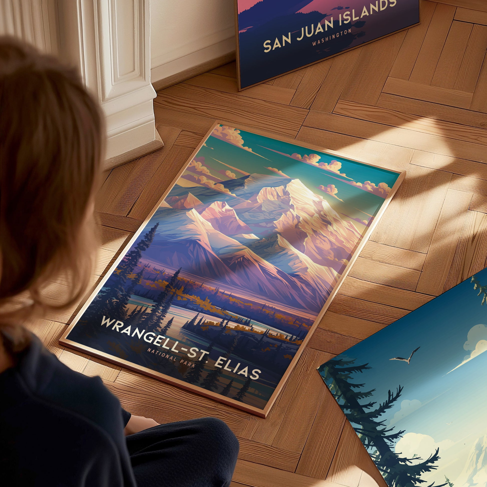 A person views a vibrant Wrangell-St. Elias National Park poster on a wooden floor, featuring dramatic mountain peaks and a scenic landscape. Another poster is partially visible nearby.