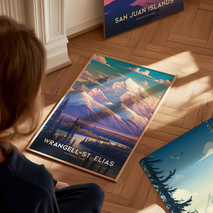 A person views a vibrant Wrangell-St. Elias National Park poster on a wooden floor, featuring dramatic mountain peaks and a scenic landscape. Another poster is partially visible nearby.