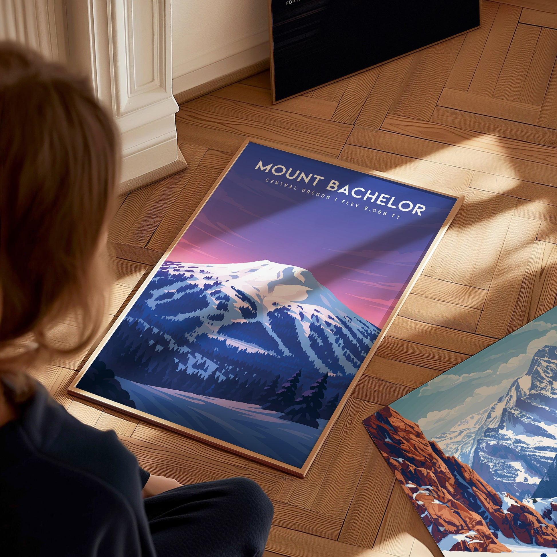 A person sits on a wooden floor, gazing at a vibrant art print featuring Mount Bachelor, showcasing snowy peaks and a colorful sunset sky. Nearby, another art print of a mountainous landscape lies partially visible on the floor.