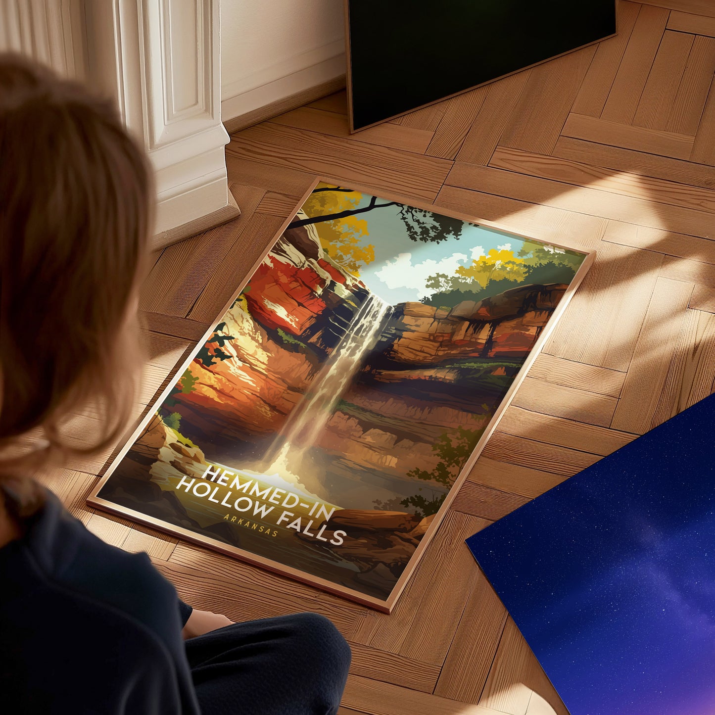 A child sits on a wooden floor, gazing at a vibrant poster depicting Hemmed-In Hollow Falls in Arkansas, featuring a waterfall cascading over colorful cliffs surrounded by lush greenery. Nearby, a starry sky print is partially visible.