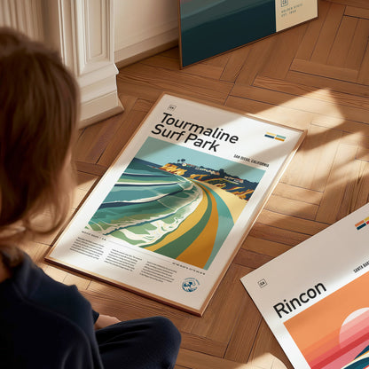 A person is sitting on a wooden floor, looking at a framed poster titled Tourmaline Surf Park featuring a colorful beach scene. Other posters are partially visible nearby.