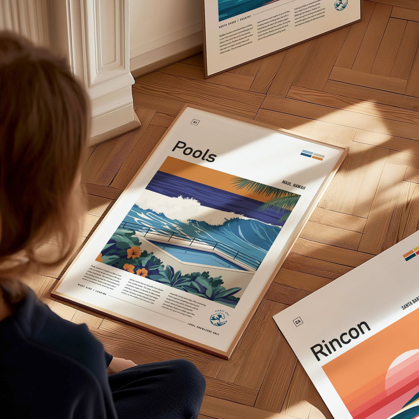 A person is sitting on a wooden floor, looking at a framed poster titled Pools, depicting a vibrant surf scene with waves and tropical foliage.