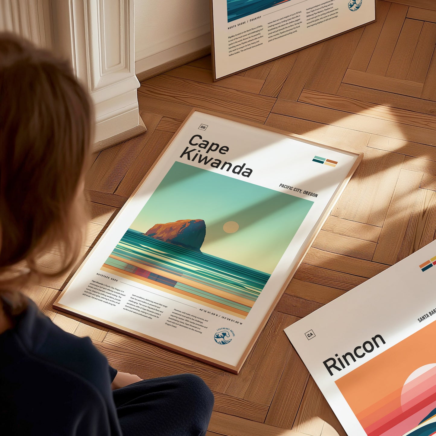 A person sits on a wooden floor, looking at a Cape Kiwanda poster featuring a stylized ocean scene with a rock formation and sunset. Other posters are partially visible nearby.