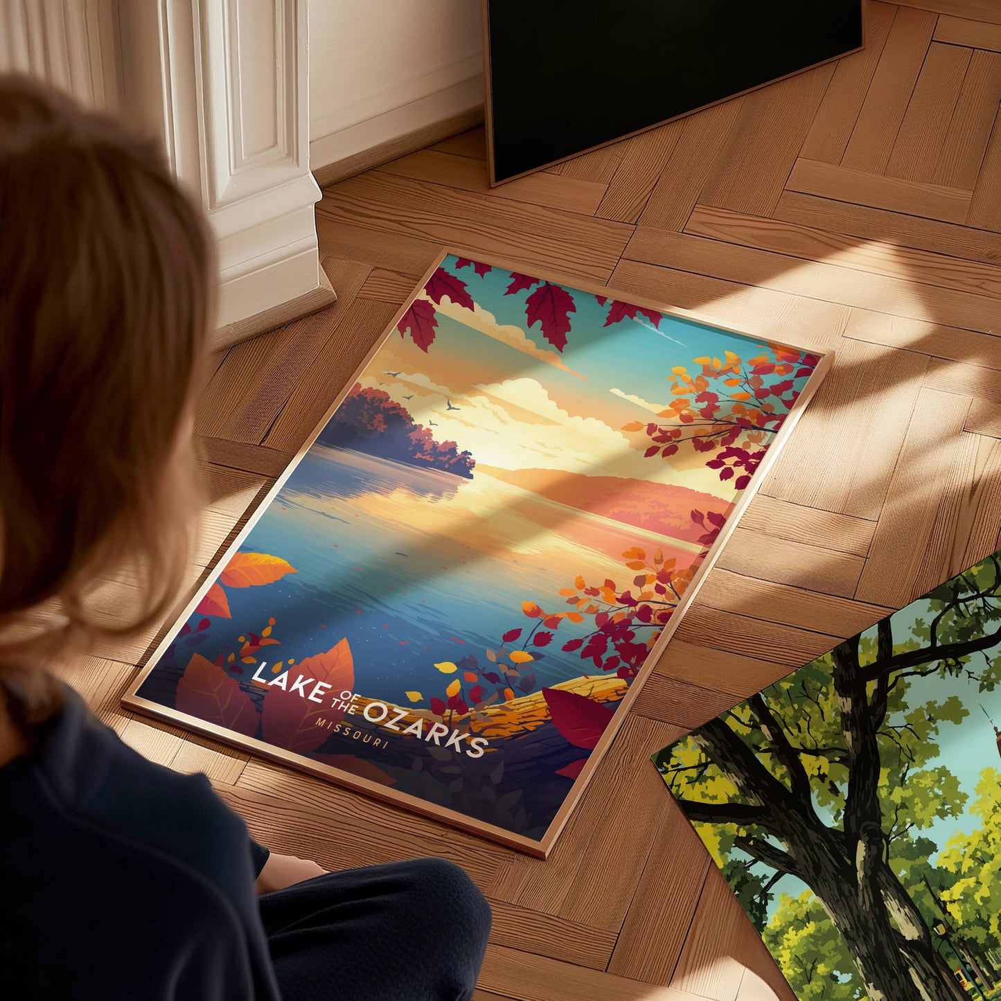 A person sits on a wooden floor, gazing at a colorful poster depicting a serene lake scene at Lake of the Ozarks, Missouri. The artwork features vibrant autumn leaves and a warm sunset over the water, creating a tranquil atmosphere.