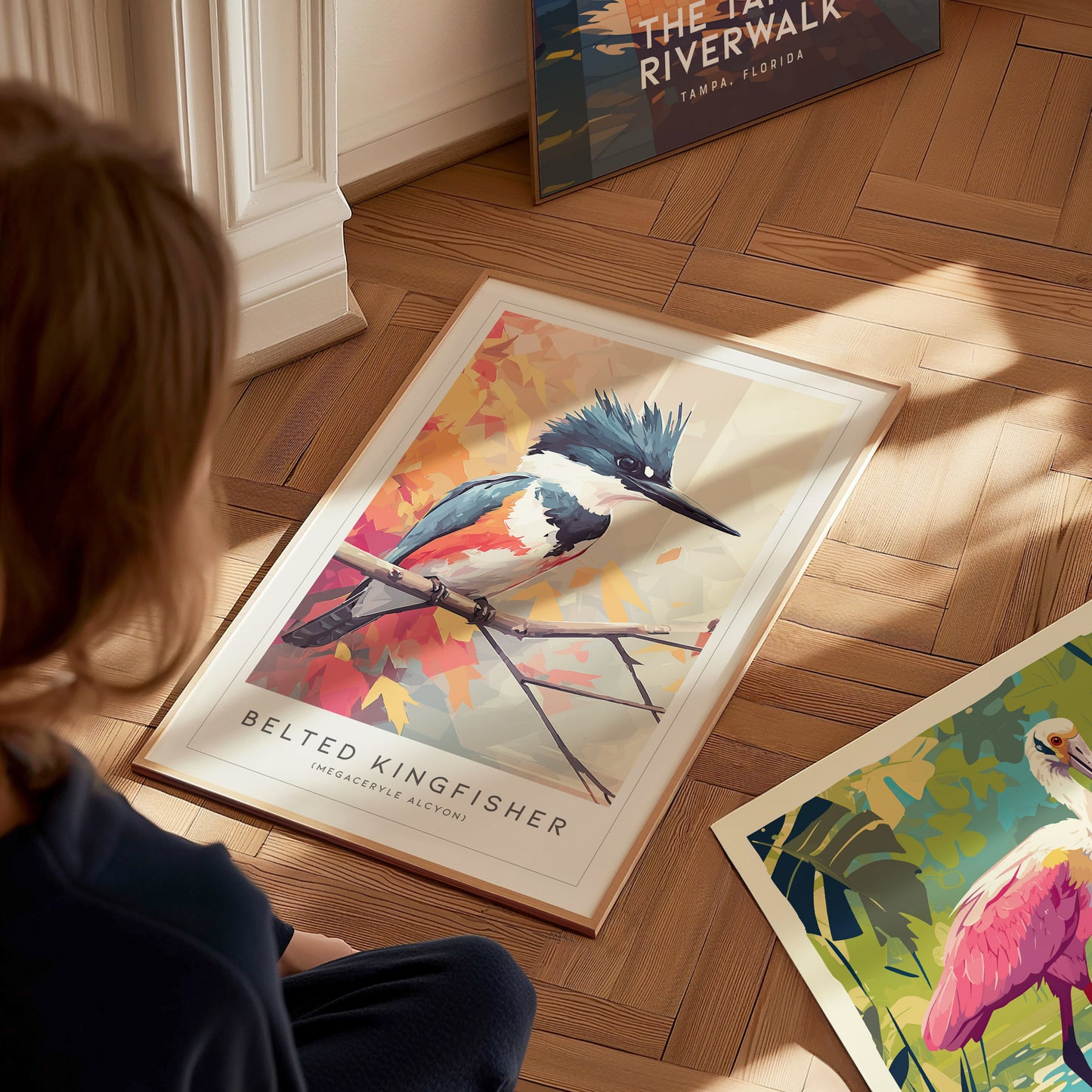 A child sits on a wooden floor, admiring a framed art print of a Belted Kingfisher perched on a branch with colorful autumn leaves in the background. Other art prints are visible nearby, showcasing vibrant nature scenes.