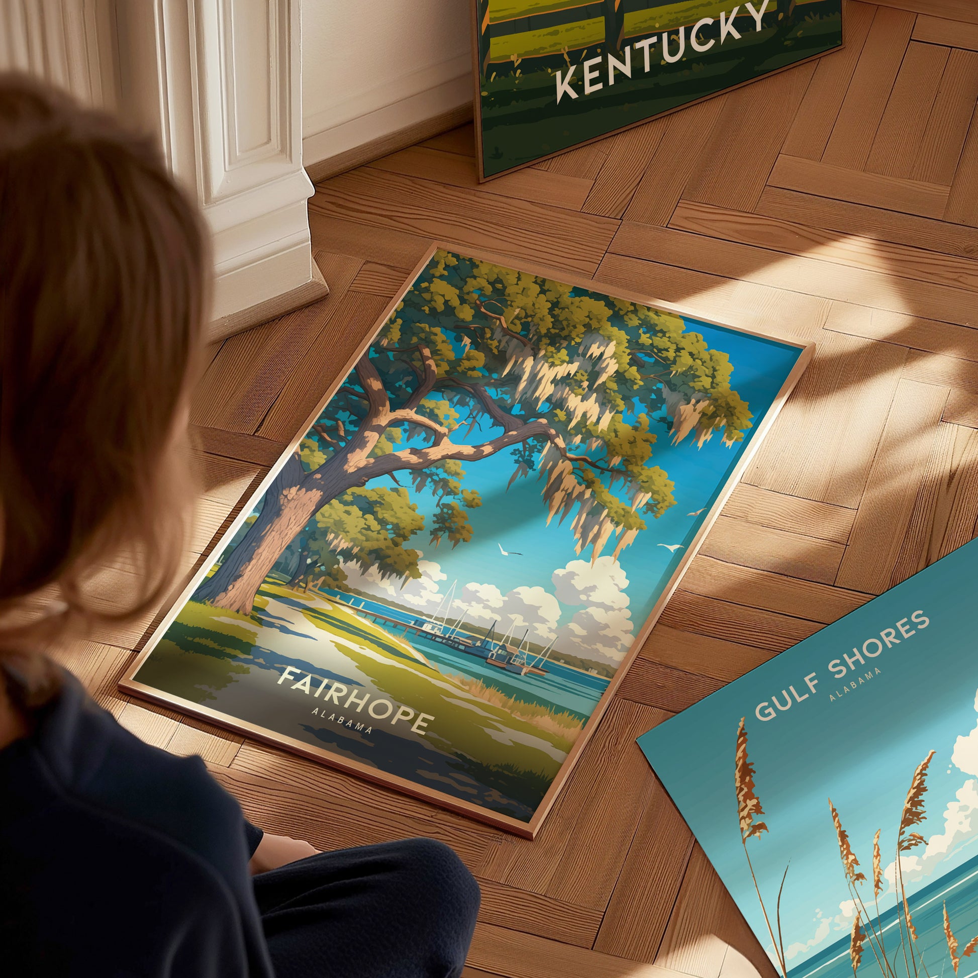 A person gazes at a vibrant Fairhope, Alabama poster on a wooden floor, featuring a scenic view of a tree by the water. Other posters are partially visible.