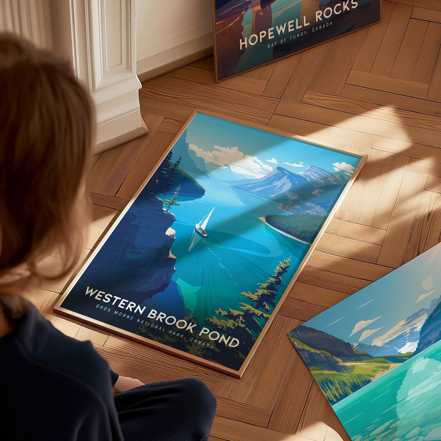 A person looks at a vibrant poster on the floor depicting Western Brook Pond with a sailboat on turquoise waters, surrounded by steep cliffs. Another poster is partially visible nearby.