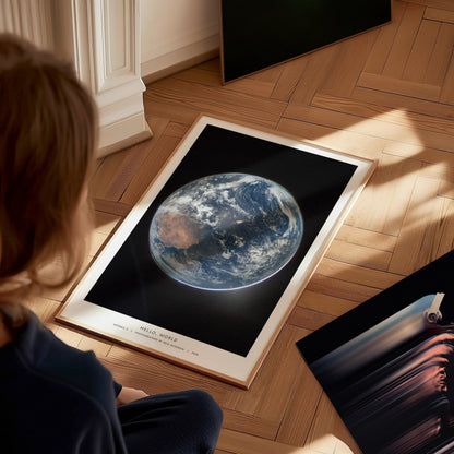 A child sits on a wooden floor, gazing at a framed poster featuring a stunning view of Earth from space, with vibrant colors highlighting the planets surface.