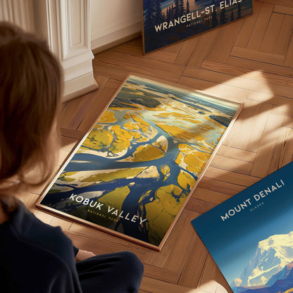 A person sits on a wooden floor, looking at a vibrant Kobuk Valley National Park poster depicting abstract landscapes. Nearby are Wrangell-St. Elias and Mount Denali posters.