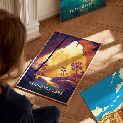 A person looks at a colorful Mammoth Cave National Park poster on a wooden floor, showcasing a vibrant cave scene with sunlight and trees. Another poster is partially visible nearby.