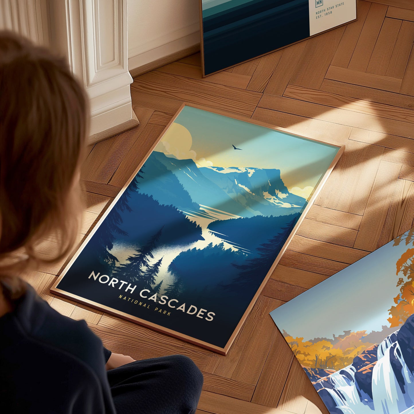 A person looks at a North Cascades National Park poster on a wooden floor, depicting mountains, a lake, and trees in a stylized design. Sunlight casts shadows across the scene.