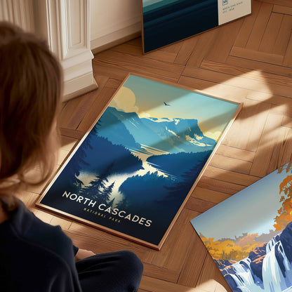 A person looks at a North Cascades National Park poster on a wooden floor, depicting mountains, a lake, and trees in a stylized design. Sunlight casts shadows across the scene.