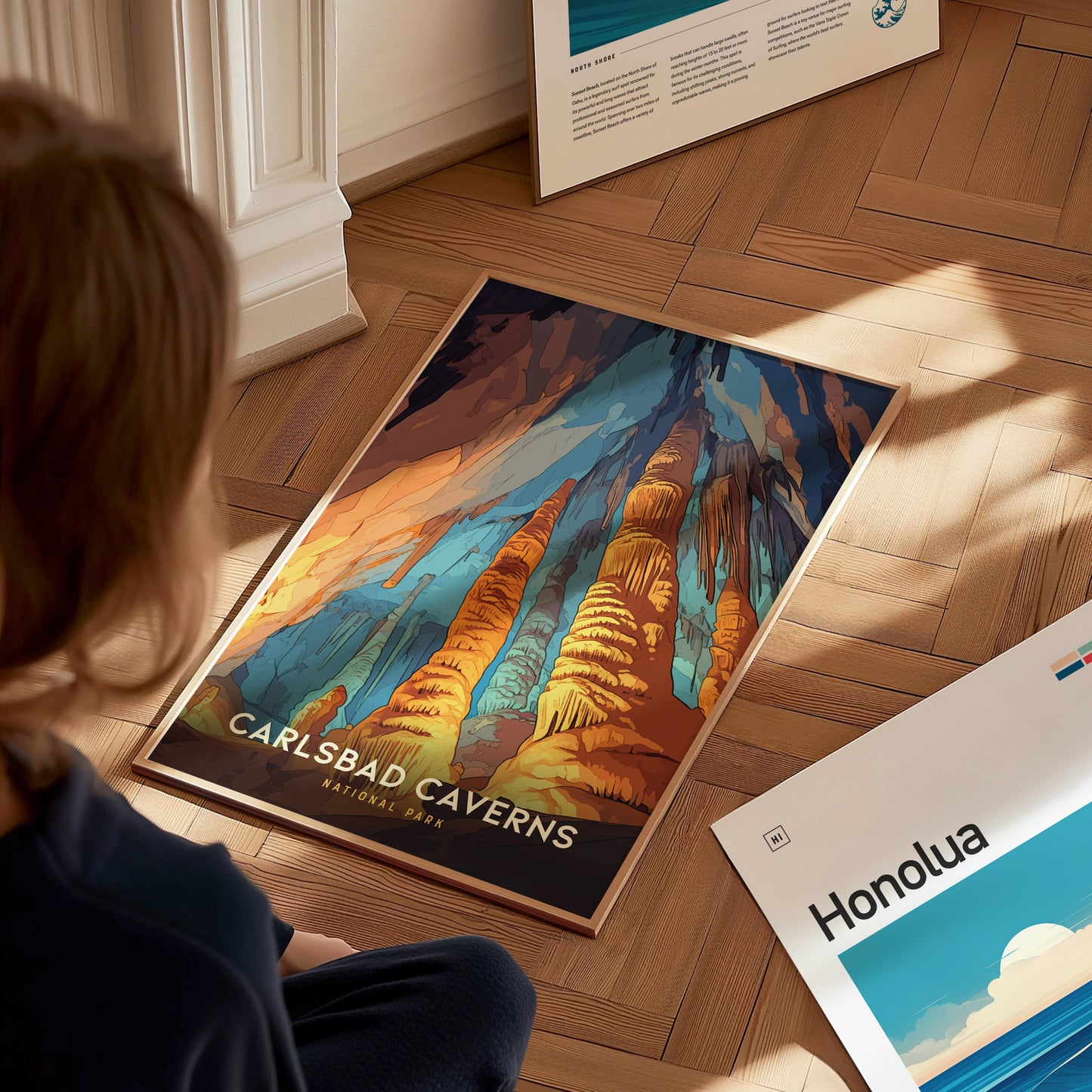 A person sits on a wooden floor, looking at a colorful Carlsbad Caverns National Park poster featuring vibrant rock formations. Another poster labeled Honolua is partially visible nearby.