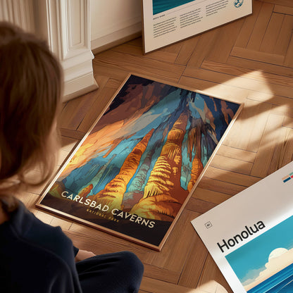 A person sits on a wooden floor, looking at a colorful Carlsbad Caverns National Park poster featuring vibrant rock formations. Another poster labeled Honolua is partially visible nearby.