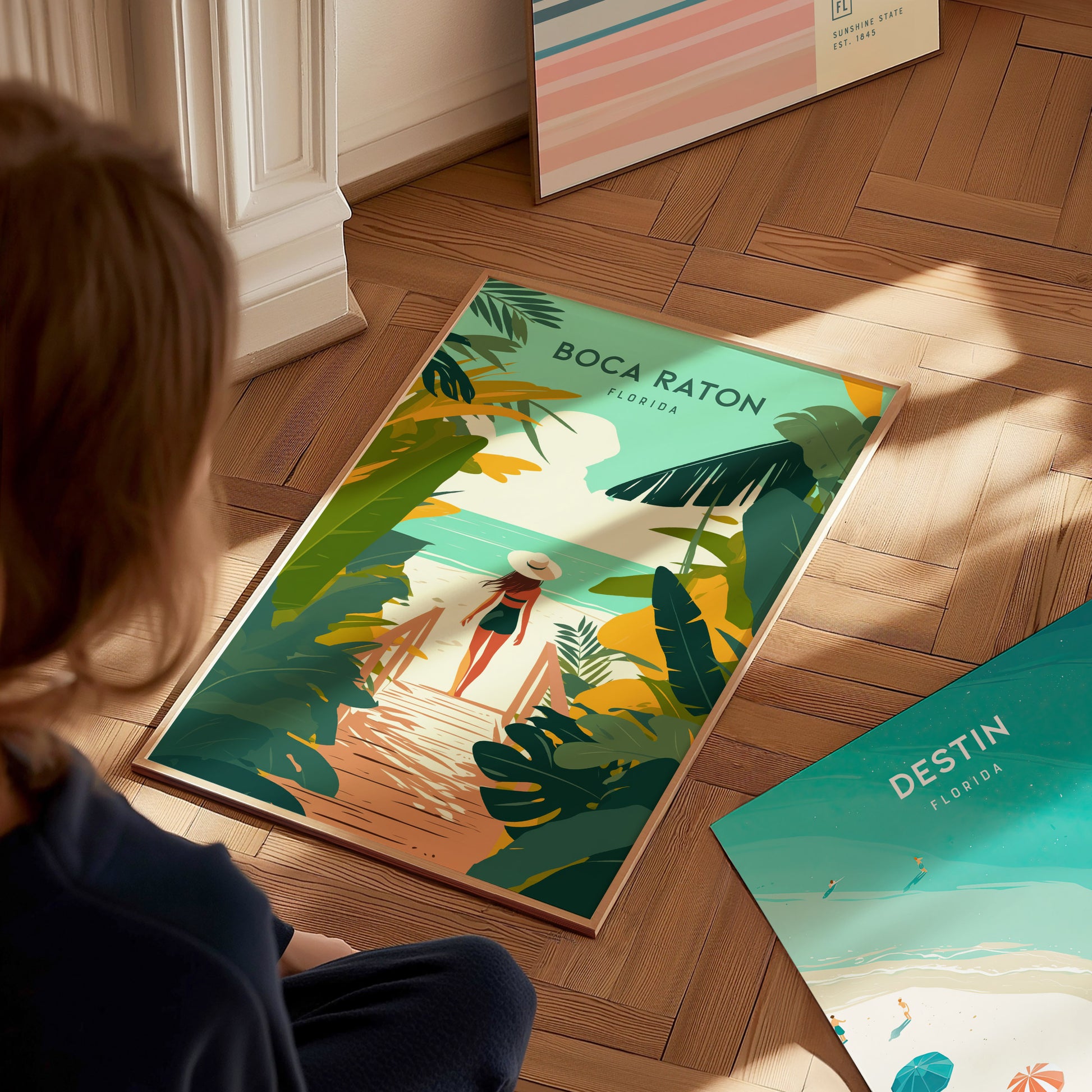 A person looks at a colorful Boca Raton travel print featuring a beach path lined with tropical plants, leading to the ocean.
