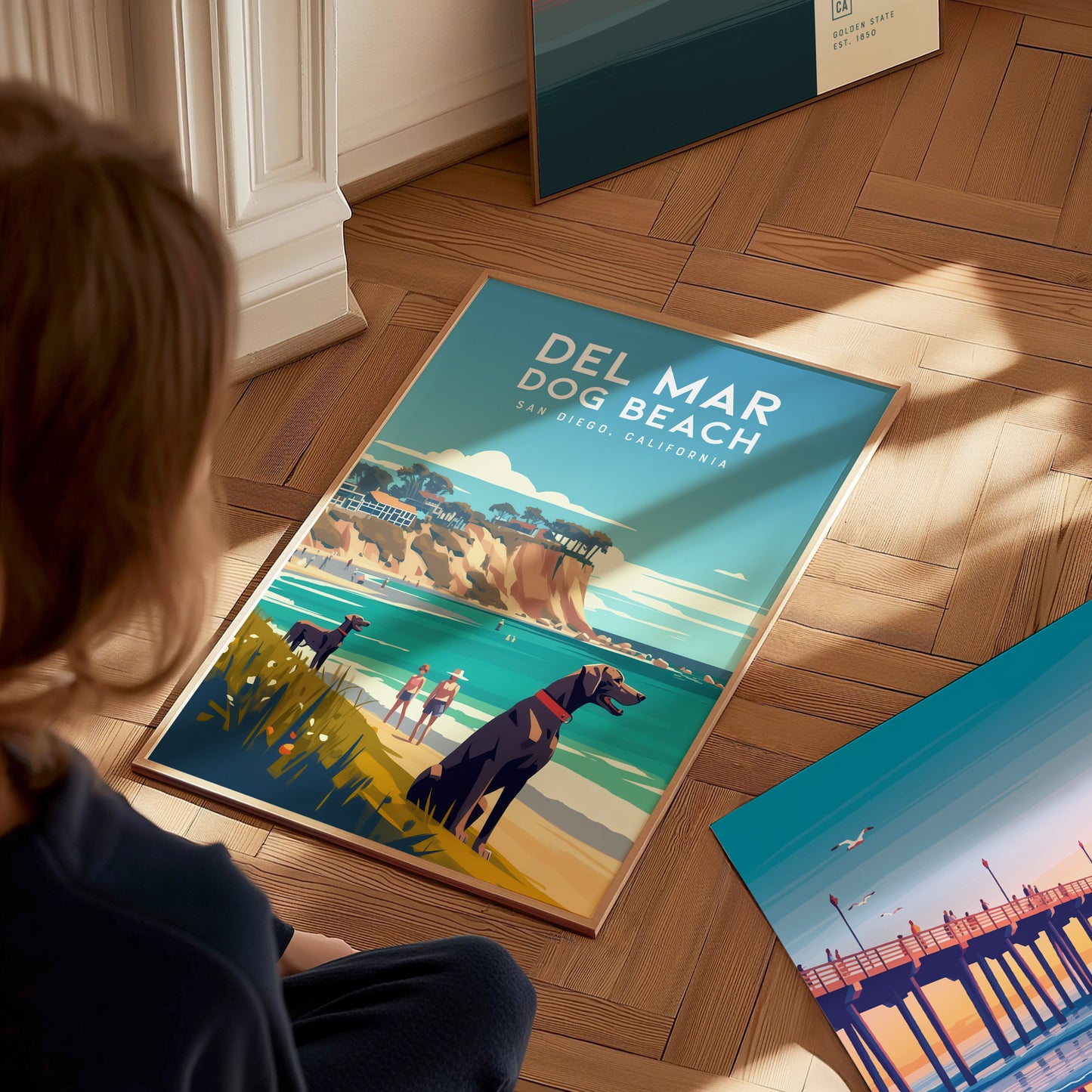 A person sits on a wooden floor, looking at a Del Mar Dog Beach poster featuring dogs, cliffs, and the ocean. Another coastal-themed poster is nearby, leaning against the wall.