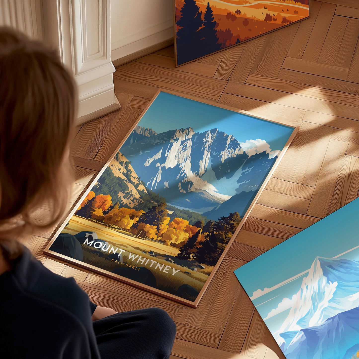 A person sits on a wooden floor, looking at a Mount Whitney poster featuring vibrant autumn trees and towering granite peaks under a bright blue sky.