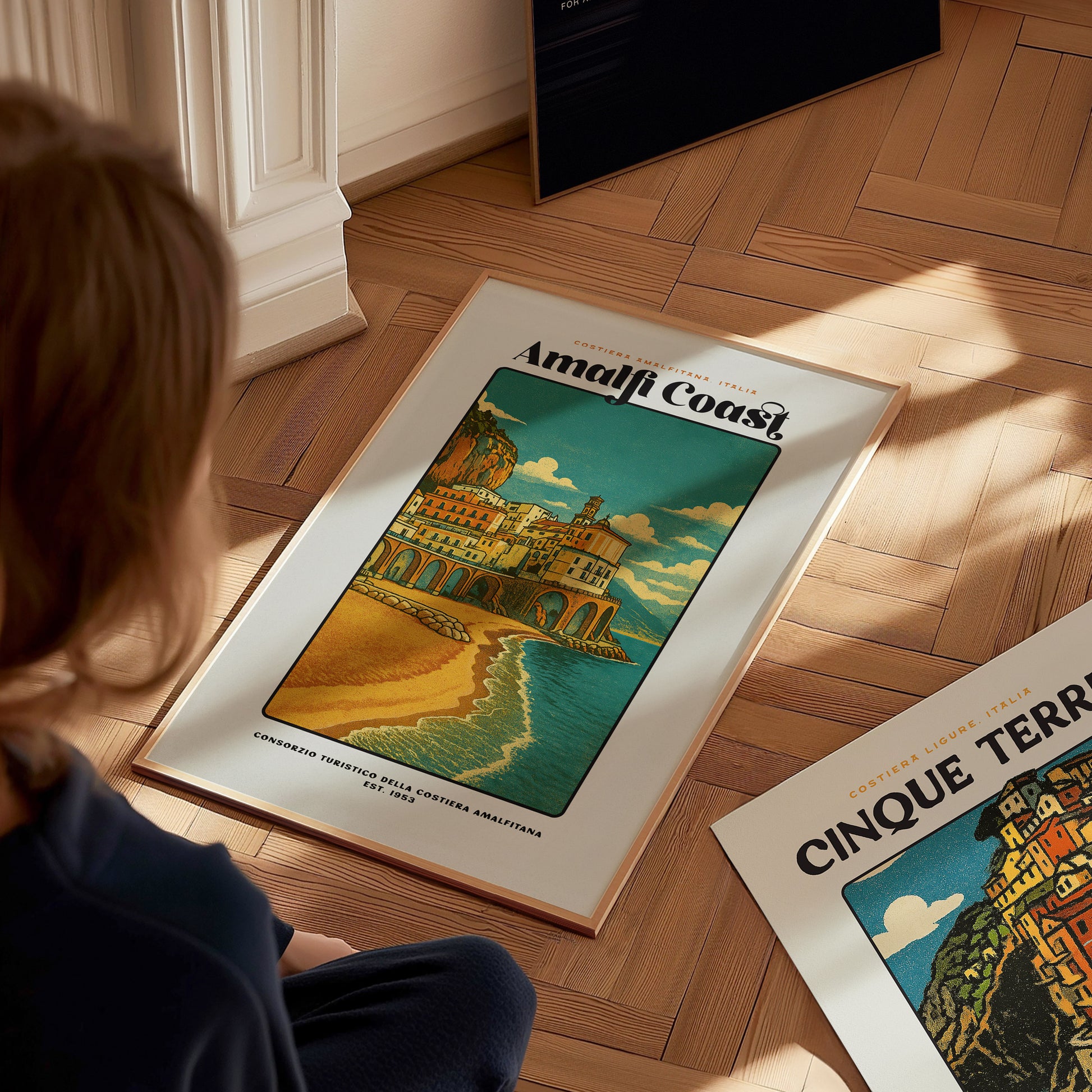 Person looking at a framed poster of the Amalfi Coast on a wooden floor.