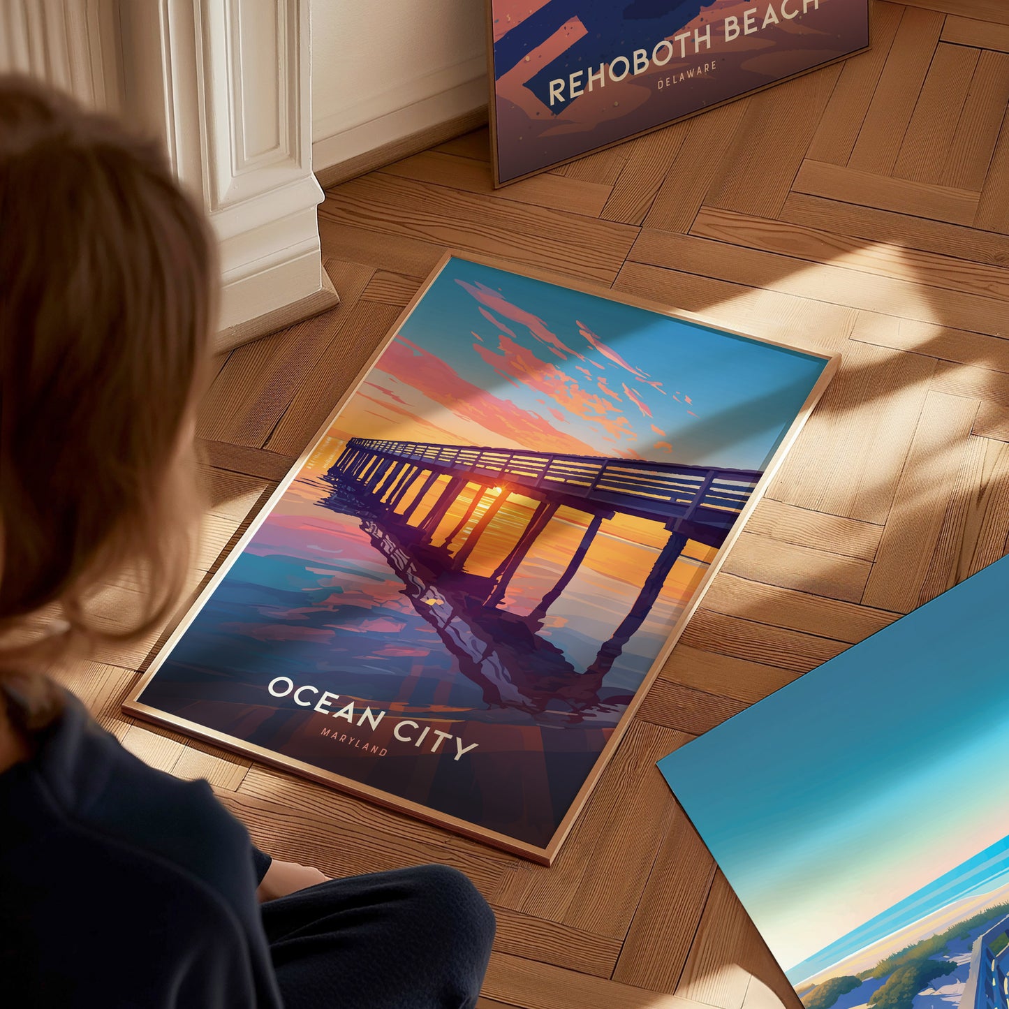 A person observes a vibrant Ocean City Maryland poster featuring a sunset view of a boardwalk pier. The art print rests on a wooden floor beside another beach-themed poster.